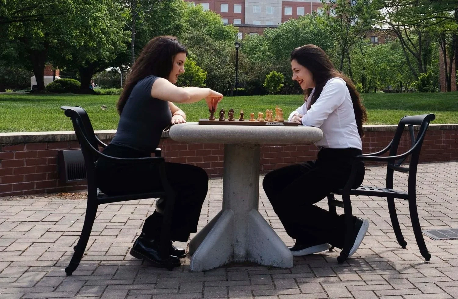 Two women playing chess outdoors at a round stone table in a park with green grass, trees, and buildings in the background.