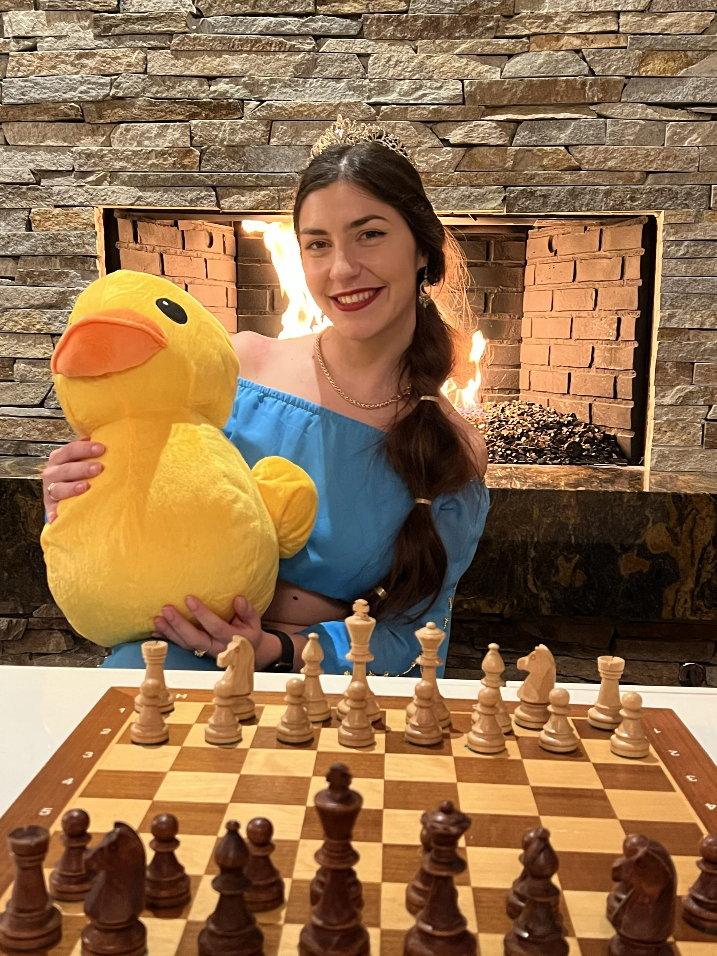 A woman wearing a blue dress, a tiara, and earrings, holding a large yellow plush duck, sitting in front of a fireplace with a chess set on a board in the foreground.