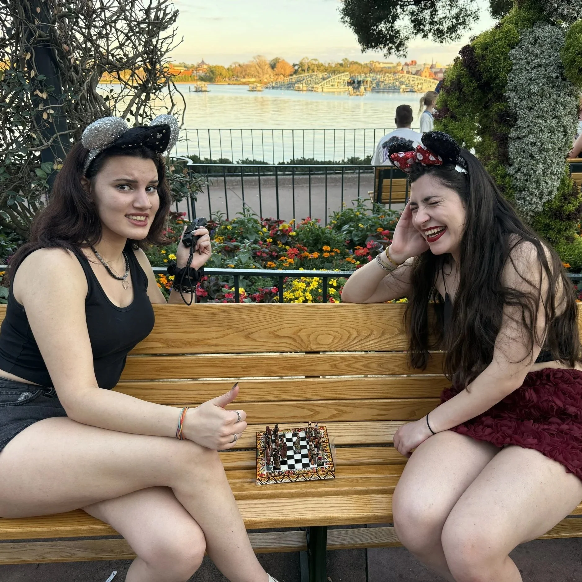 Two women with Minnie Mouse ears headbands playing chess outdoors on a wooden bench, lake and bridge in the background.