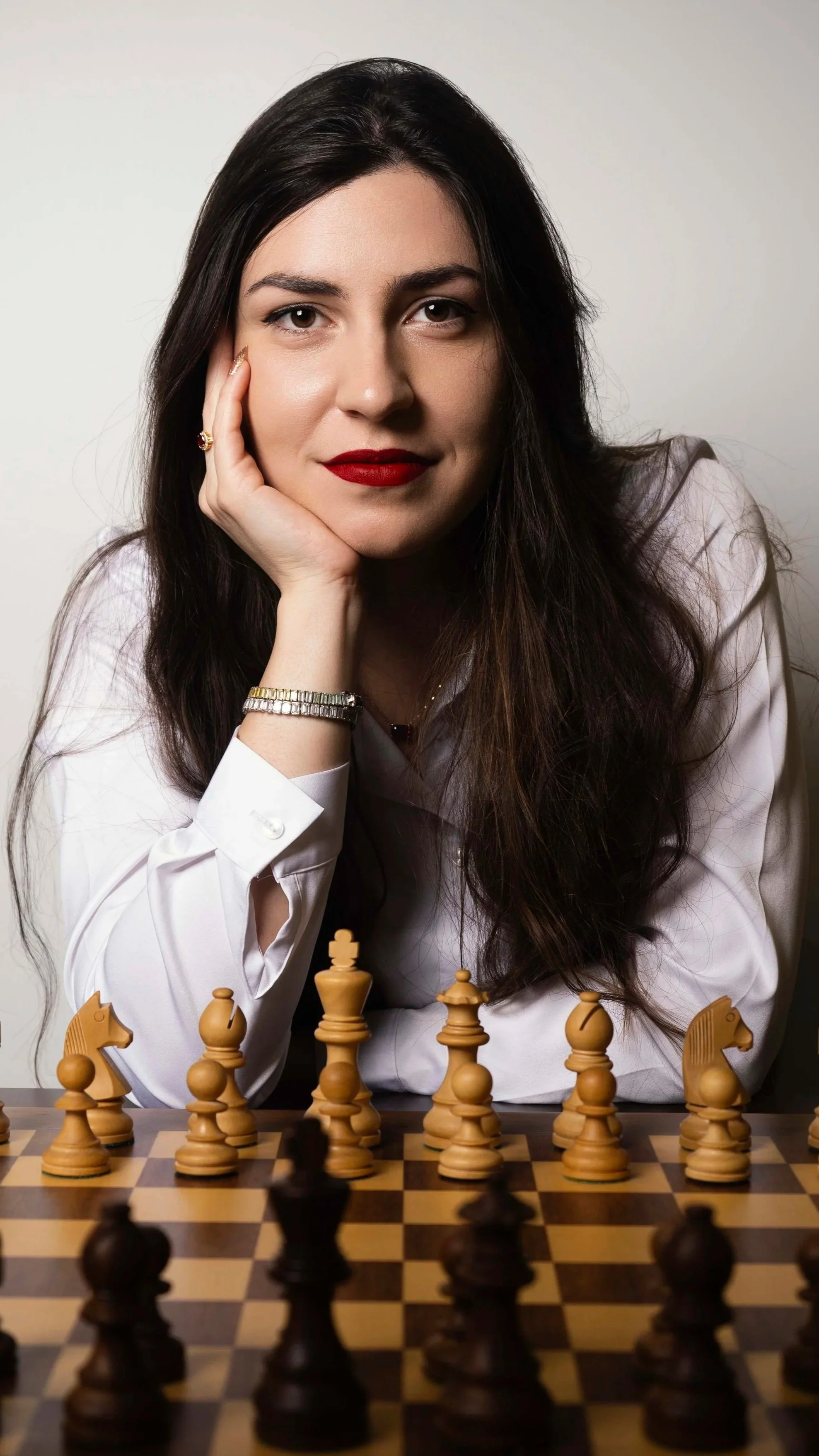 A woman with long dark hair and red lipstick sitting at a chessboard with chess pieces in front of her, resting her chin on her hand.
