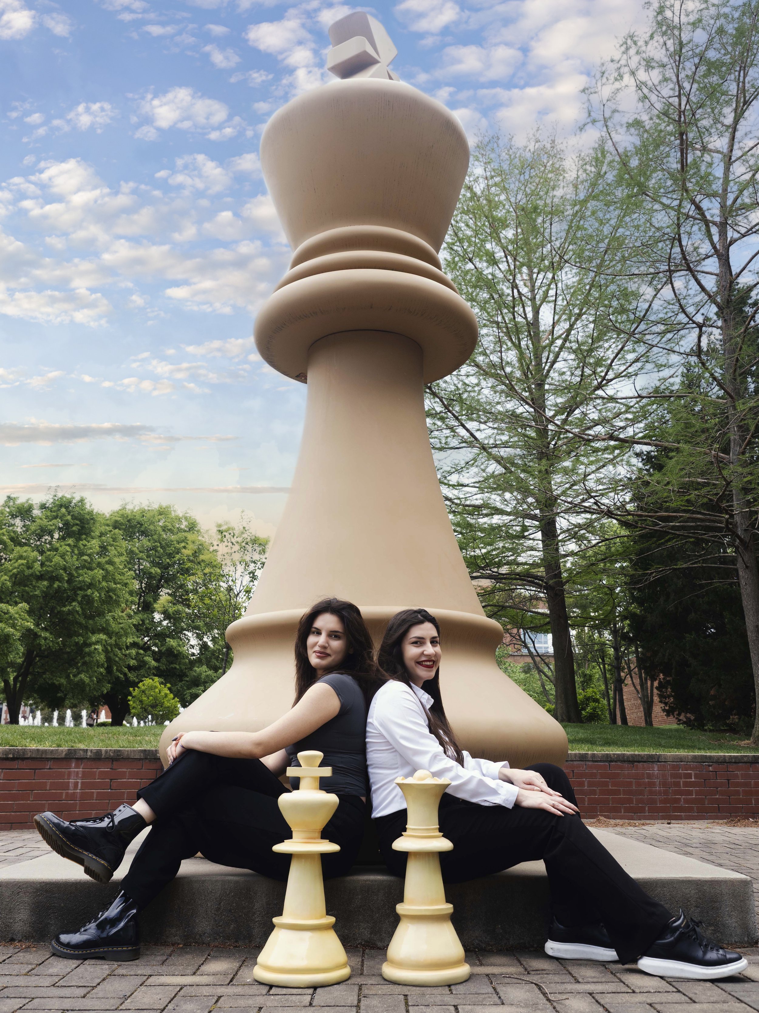 Two women sitting back-to-back in front of a large chess pawn sculpture outdoors, with trees and a cloudy sky in the background.