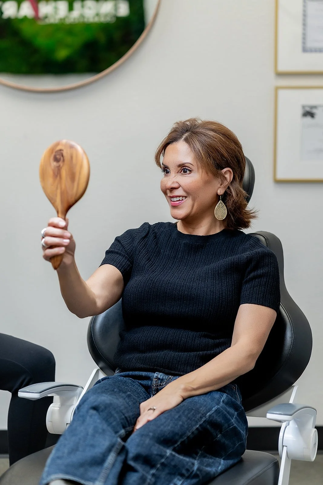 A woman with short brown hair and earrings sitting in a black chair, smiling, holding a wooden mirror, in an office setting.