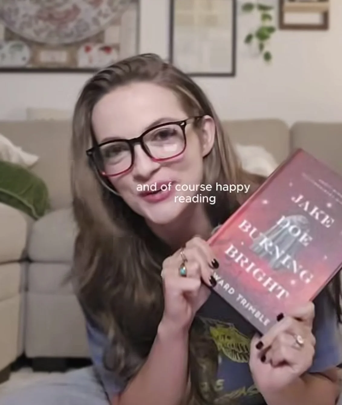 A woman with long brown hair and glasses smiling and holding a pink book titled 'The House in the Cerulean Sea' by TJ Klune in a cozy living room.