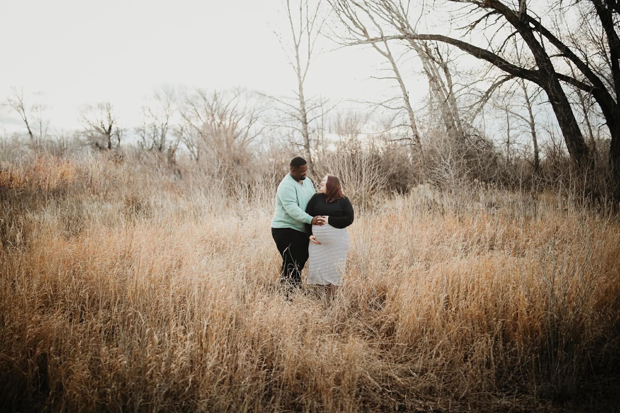 A couple standing in a field of tall, dry grass, holding hands and gazing at each other, with leafless trees in the background during overcast weather.