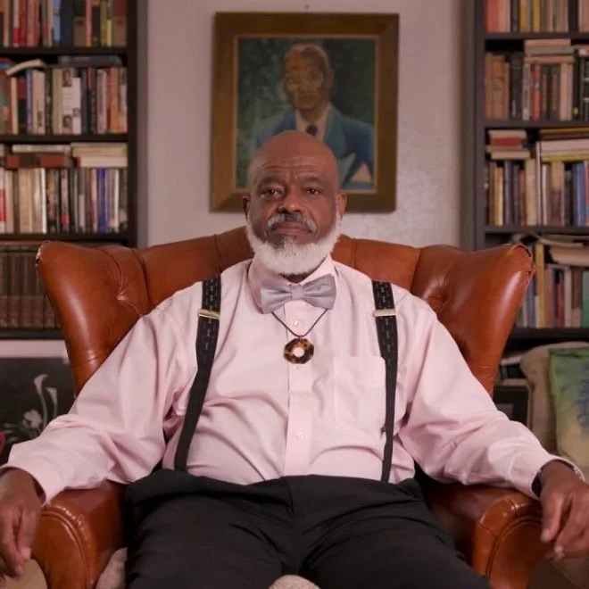 Harrison Eppright sitting in a brown leather armchair. He is wearing a white dress shirt, gray bow tie, suspenders, and a necklace. Behind him, there is a bookshelf filled with books and a portrait of a man hanging on the wall.