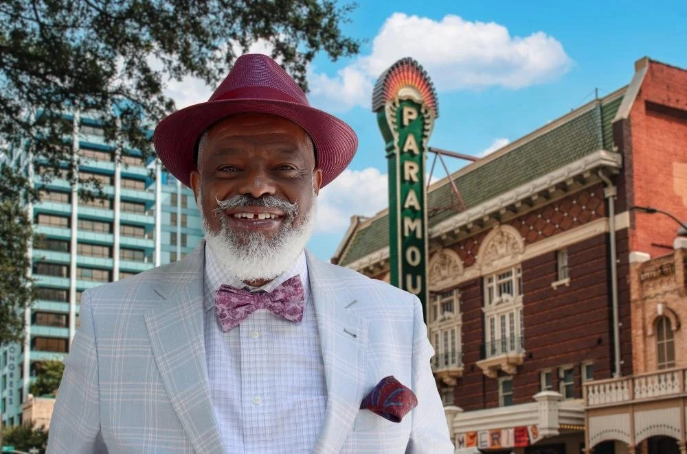 Harrison Eppright wearing a light-colored suit, a patterned bow tie, and a pocket square, standing in front of a historic building with a marquee that reads "Paramount" and a tall downtown office building in the background.