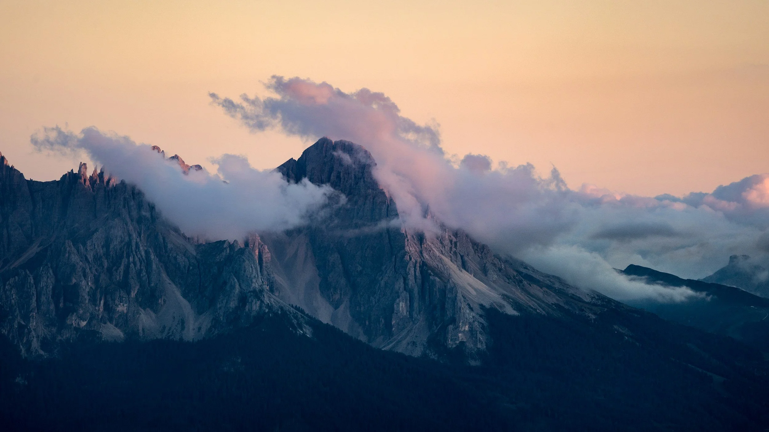 A mountain range with jagged peaks partially covered in clouds, during dusk or dawn with a light pink sky.