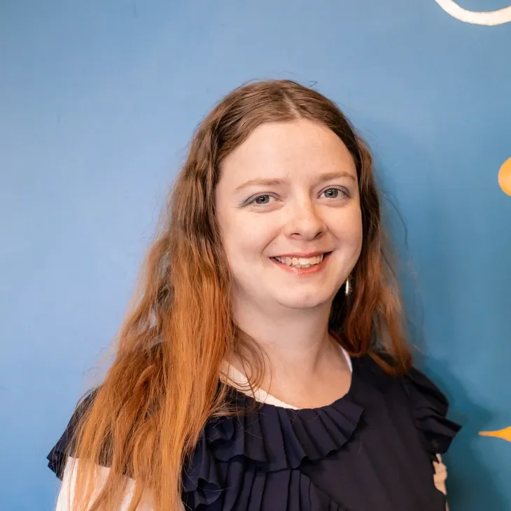 A young woman with long, wavy red hair smiling at the camera, standing in front of a blue wall with decorations.