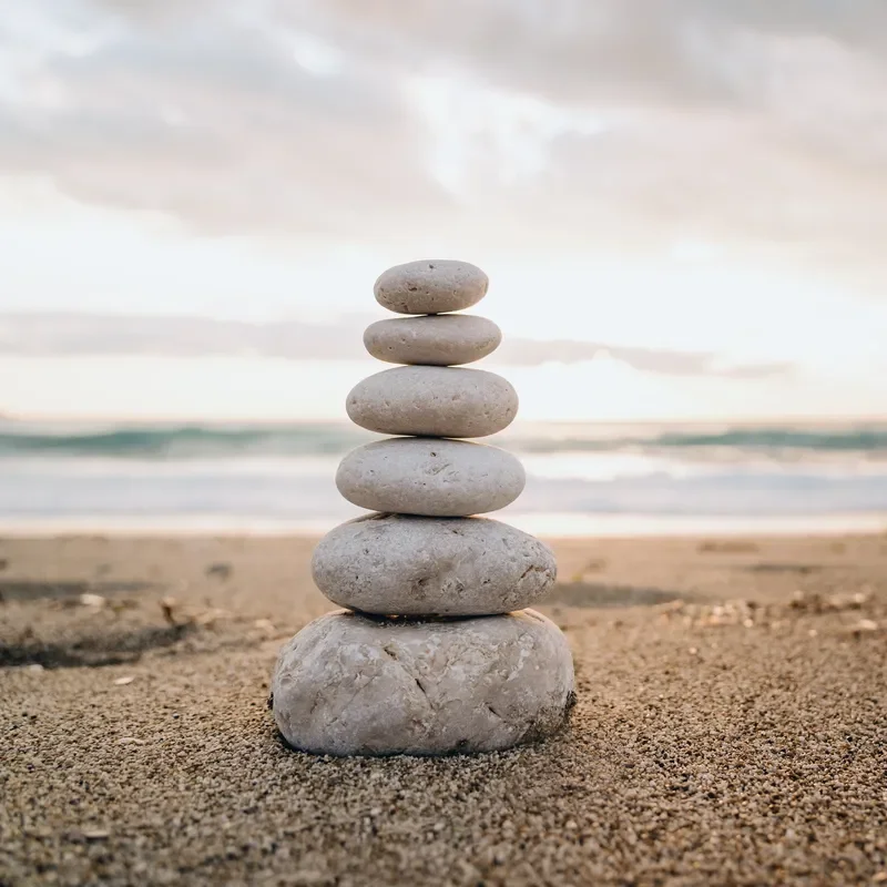 A stack of six smooth, flat stones balanced on a sandy beach with the ocean and overcast sky in the background.