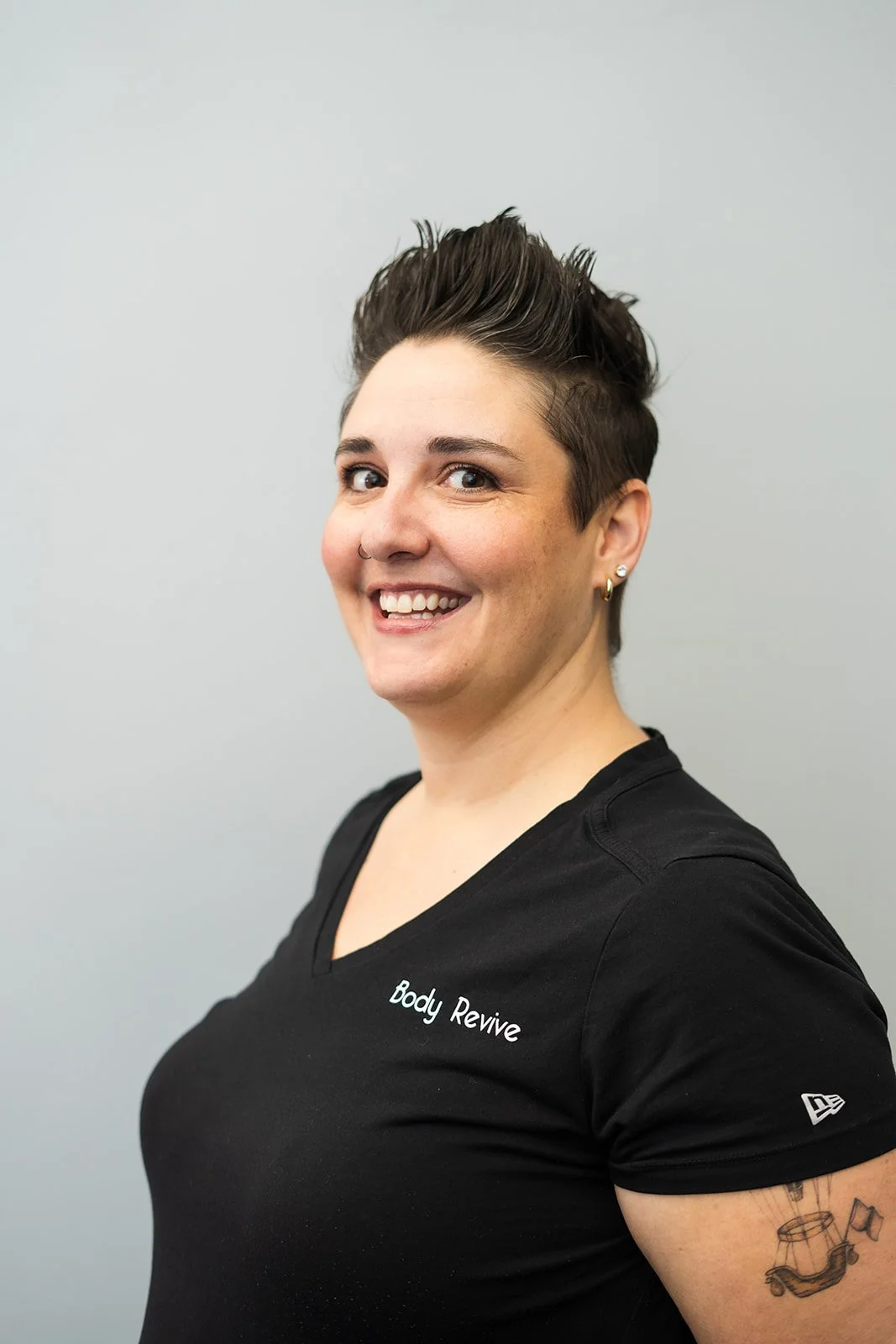A woman with short, styled dark hair, smiling at the camera, wearing a black t-shirt with 'Body Revive' written on it and earrings, standing against a plain light gray background.