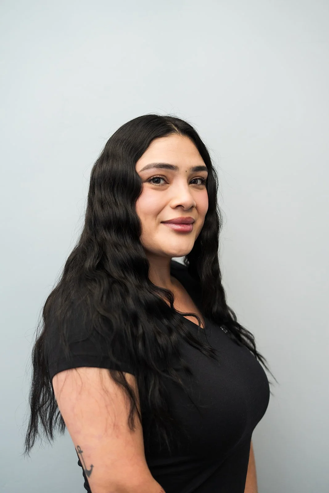 A woman with long, wavy black hair and light skin, wearing a black shirt, standing in front of a plain light-colored background.
