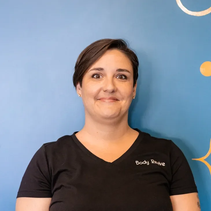 Woman with short dark hair wearing a black t-shirt with 'Body Revive' logo, standing against a blue wall with abstract orange and white design.