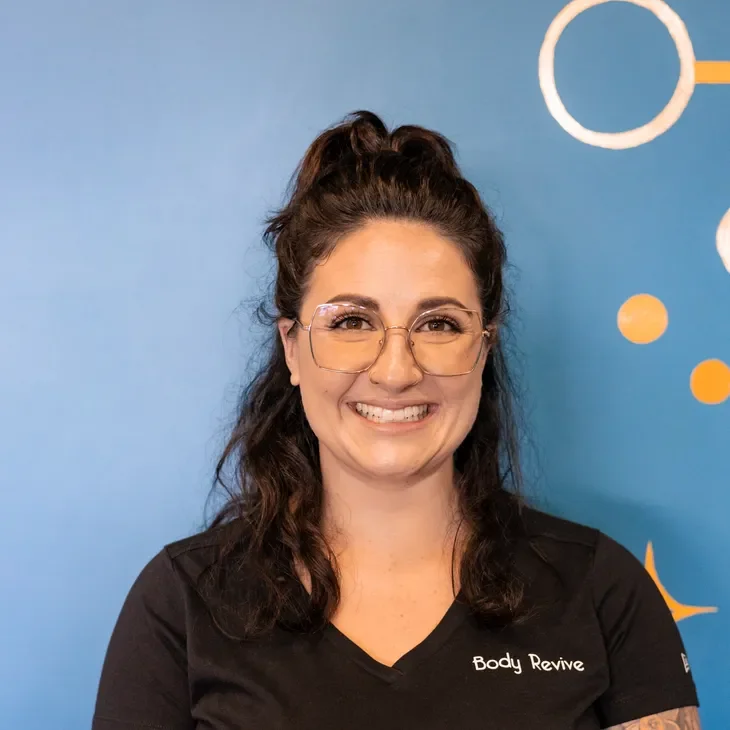 Smiling woman with glasses and dark wavy hair tied back, wearing a black shirt with 'Body Revive' logo, standing in front of a blue wall with white and orange abstract shapes.