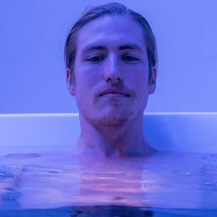 A young man with slicked-back hair relaxing in a bathtub with blue lighting.