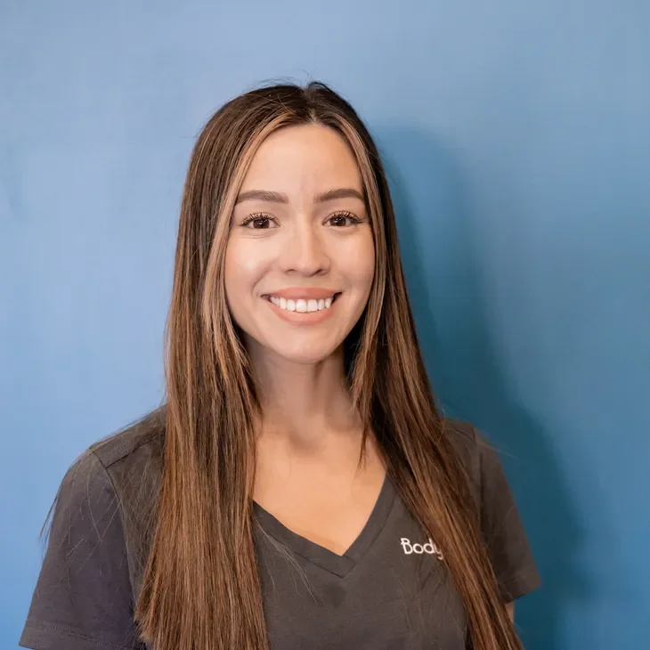A young woman with long brown hair smiling, wearing a dark gray V-neck shirt with the word "Body" visible, standing against a blue background.