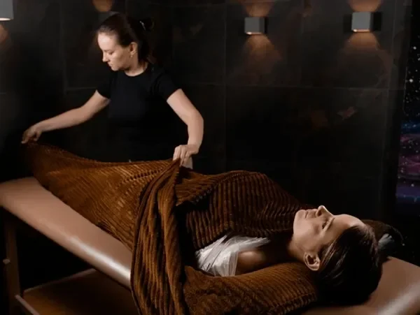 Person lying on a massage table covered with a brown blanket, receiving a massage from a therapist in a spa room.