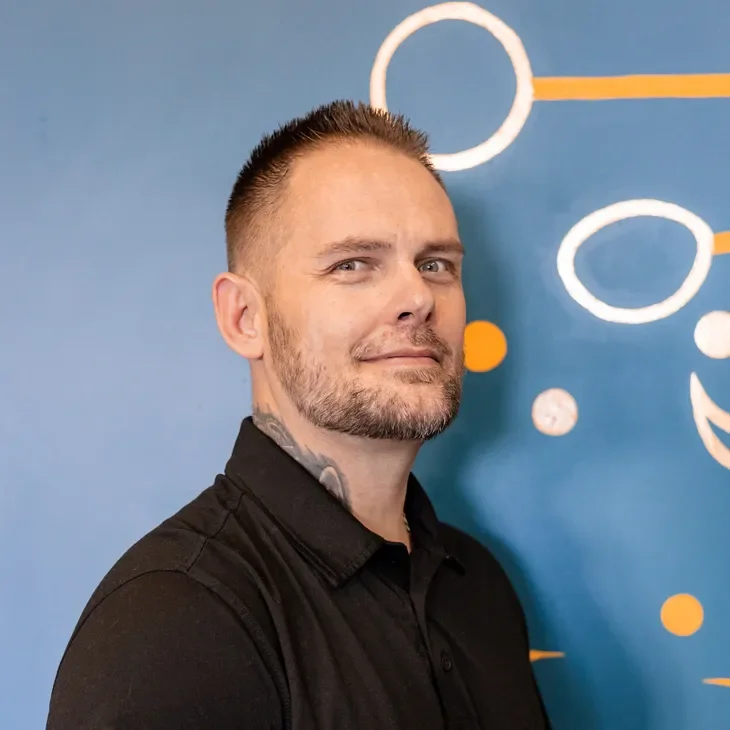 A man with short hair, beard, and a tattoo on his neck, wearing a black shirt, standing in front of a blue wall with white and orange circular designs.