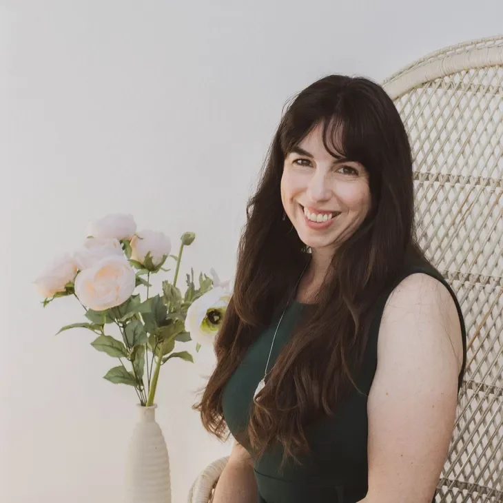 A woman with long dark hair and a green dress smiling and sitting on a wicker chair near a vase of artificial flowers.