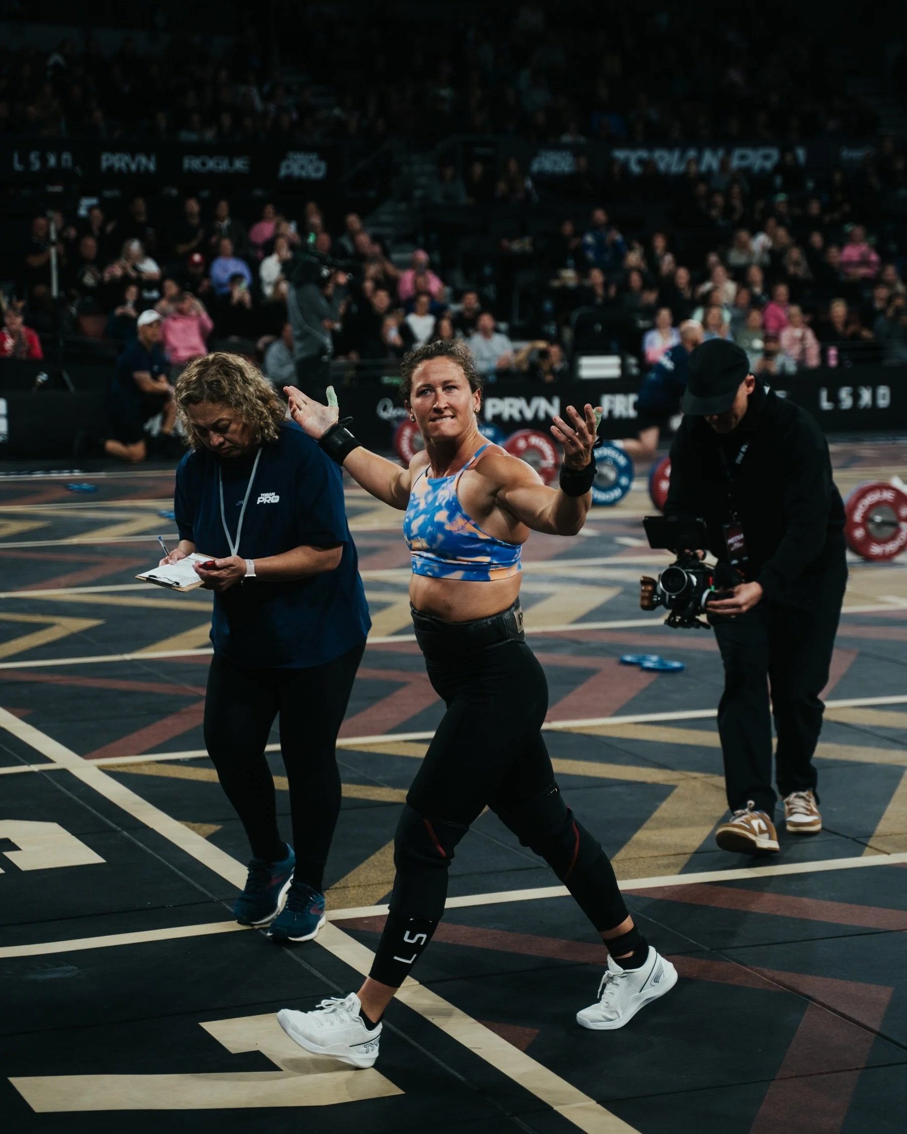 A woman in athletic gear is smiling and gesturing with her hands at a fitness event inside a large gymnasium filled with spectators. She is surrounded by event staff, with one person writing on a clipboard and another holding camera equipment.
