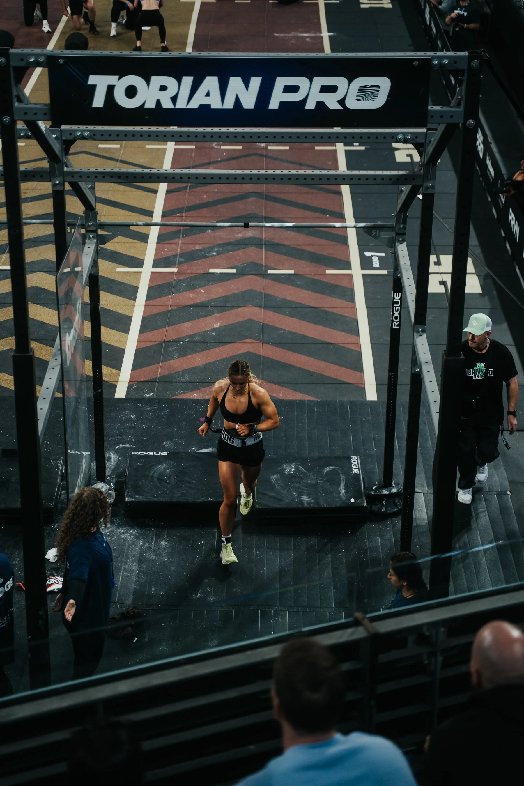 A woman crossing the finish line at a CrossFit competition, with the word 'TORIAN PRO' on the banner above. Spectators and staff are present around the event area.