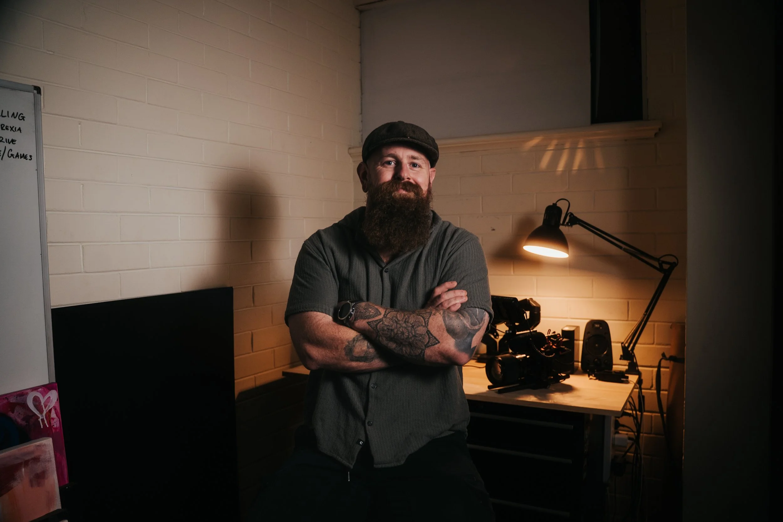 A man with a beard and tattoos on his arms, wearing a flat cap and gray shirt, leaning with arms crossed in an office or studio space, illuminated by a desk lamp, with equipment and a whiteboard in the background.