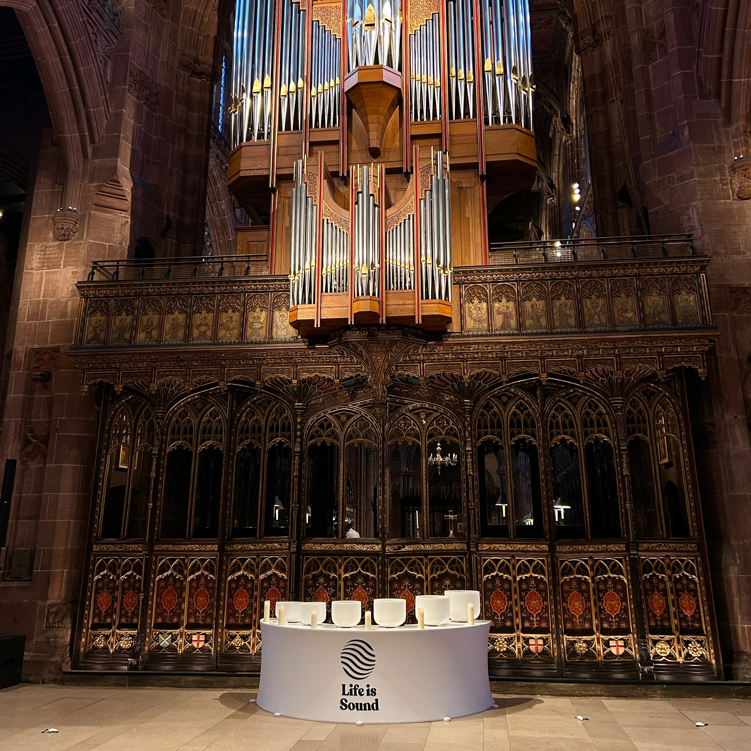 Manchester Cathedral interior with large pipe organ and ornate wooden details. There is a white table with candles, crystal singing bowls and a sign that reads "Life is Sound."