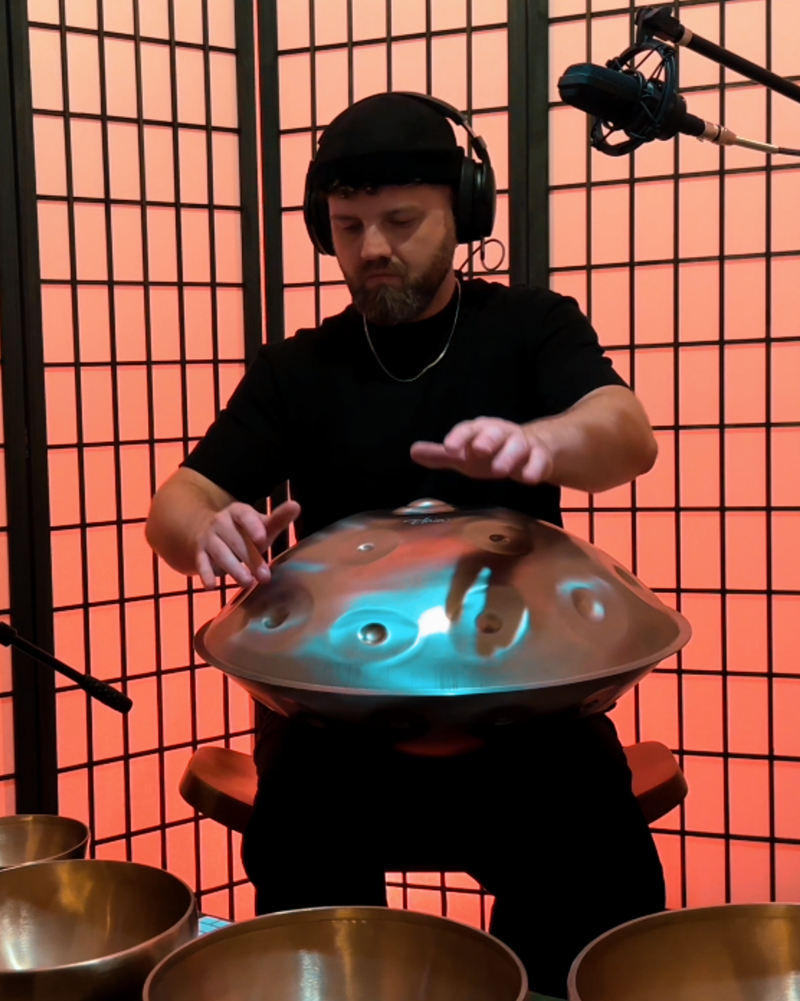 James wearing headphones and a black shirt playing a handpan, seated in front of a red grid-patterned background with other metal bowls in the foreground.
