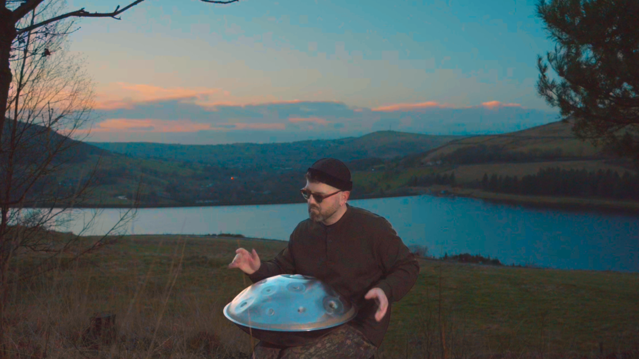 James Mayer black clothing, wearing sunglasses and a black cap, playing a handpan outdoors near a body of water during sunset with hills and trees in the background.