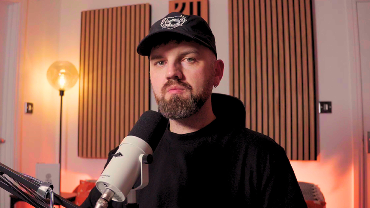 James Mayer wearing a black cap and black shirt, sits in front of a microphone in a recording studio with wooden sound panels on the wall behind him and a warm light on a lamp to the left.