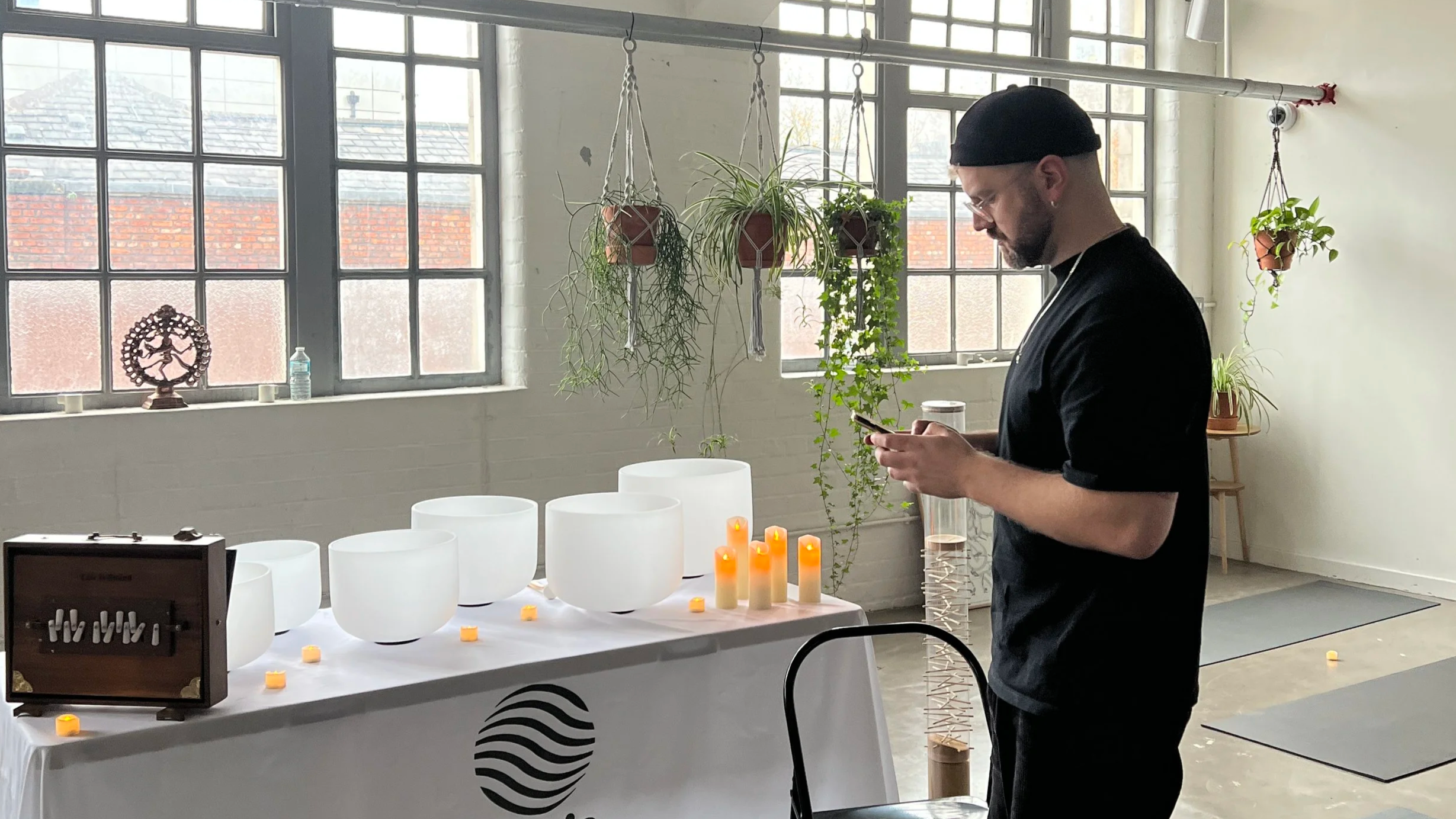 James Mayer at Space at The Mill, looking at his phone in a room with large windows and hanging plants, with white singing bowls, candles, and a table cloth with a wave logo.