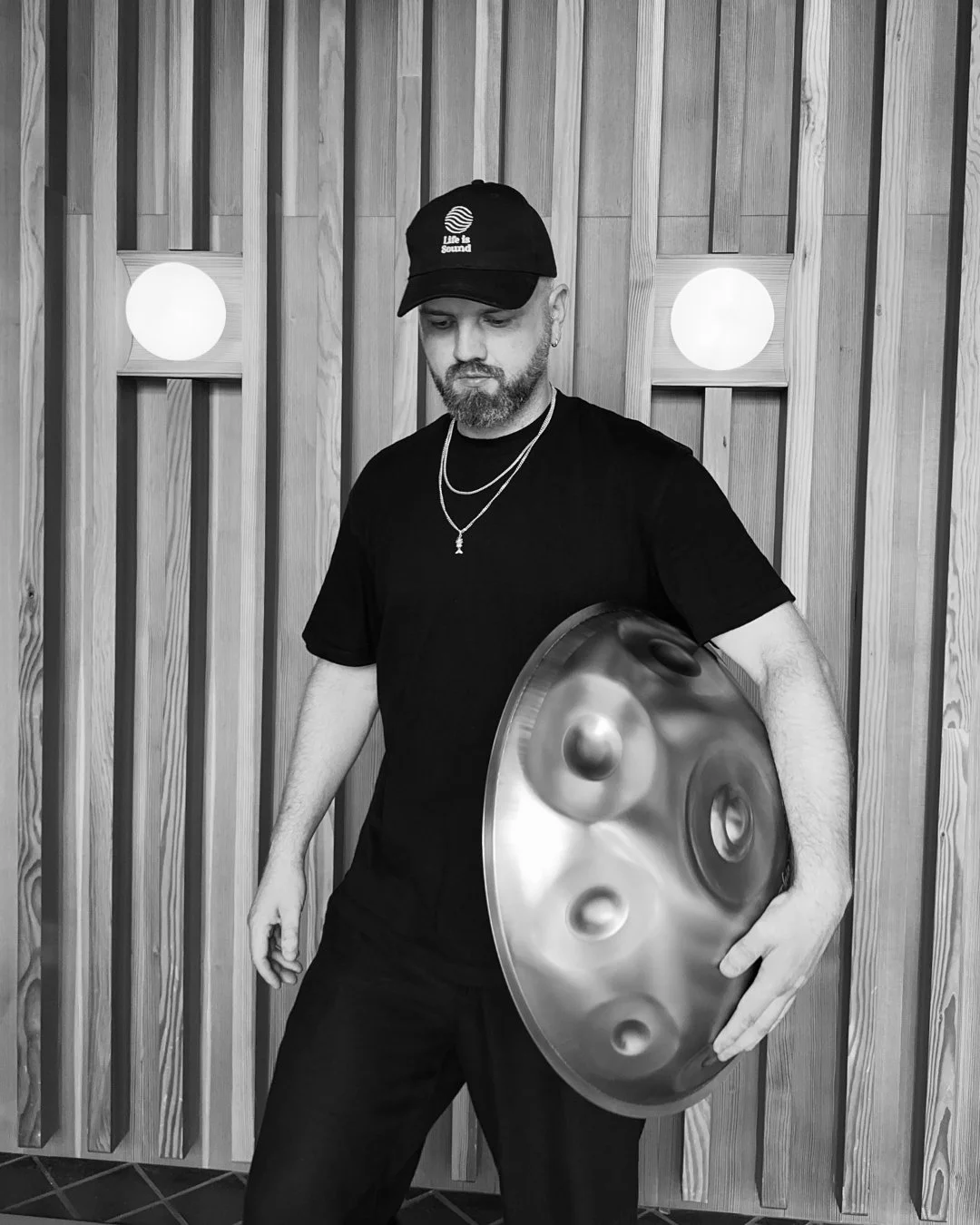 James Mayer holding a handpan, standing in front of a wooden wall with two round lights.