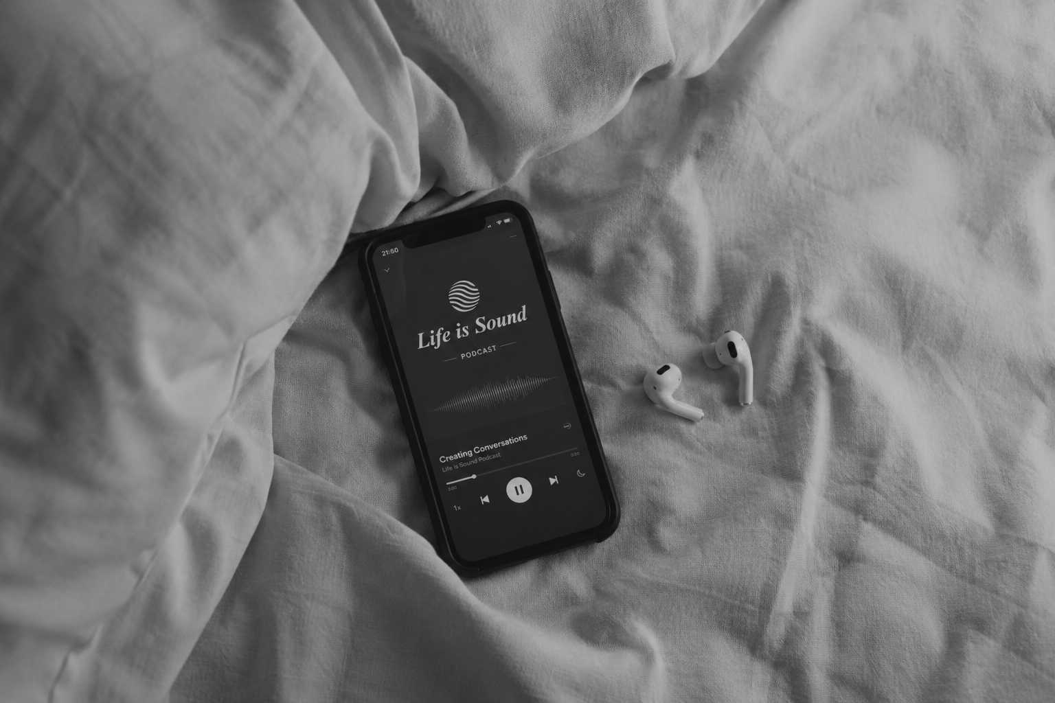 A smartphone playing a podcast called 'Life is Sound' on a bed with a pair of wireless earbuds placed next to it.