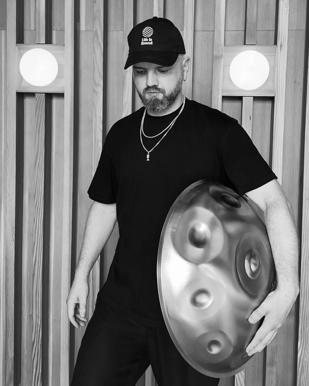 James Mayer holding handpan drum, wearing a black cap and black t-shirt, standing in front of a wooden wall with circular lights.