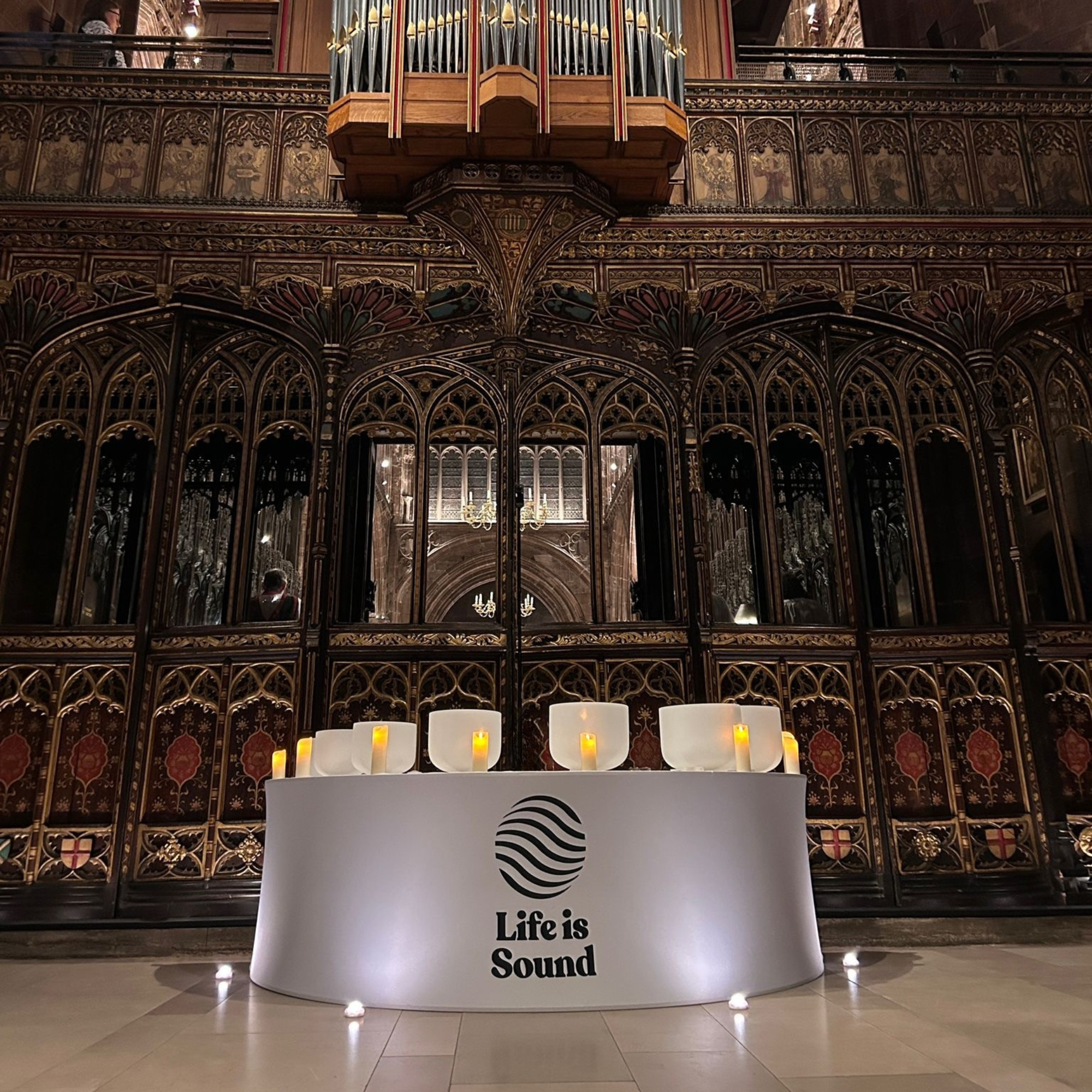 A white podium with the Life is Sound logo and the phrase "Life is Sound" in black text, illuminated by small lights around its base, in front of an ornate, gothic-architecture-style wooden structure with candles and stained glass windows in the background.