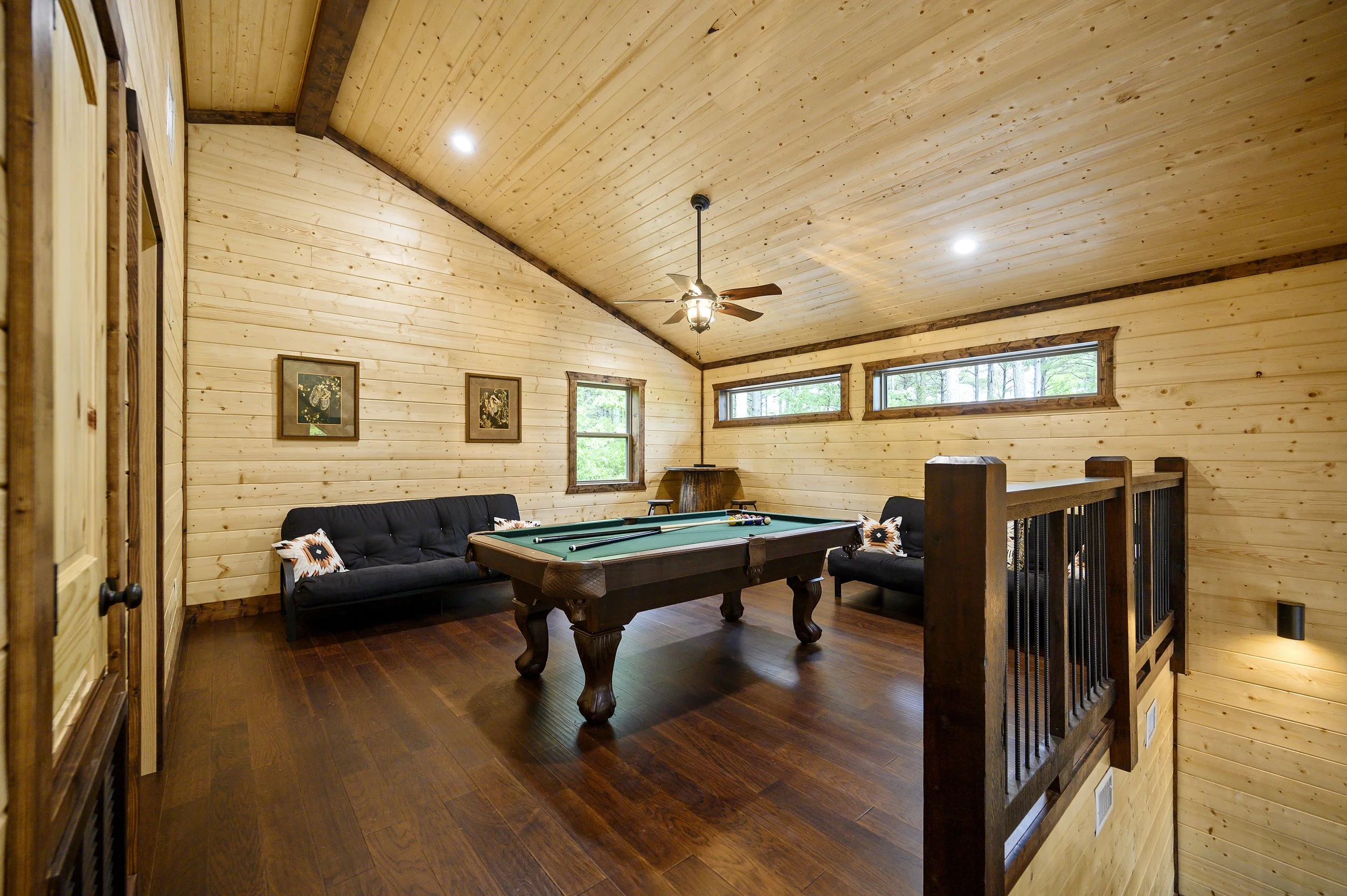 Interior of a cozy game room with wooden walls and ceiling. Features a pool table in the center, two black sofas with decorative pillows, a ceiling fan, and windows letting in natural light.