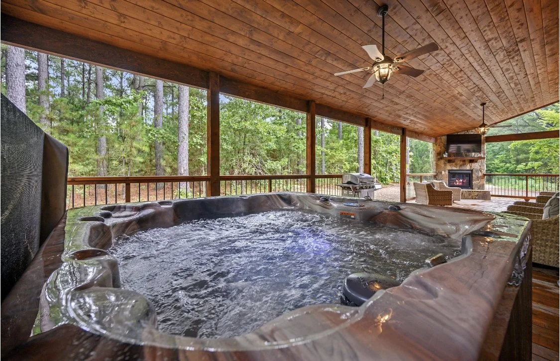 Outdoor hot tub on a covered porch with a view of a forest, featuring a fireplace and outdoor seating.
