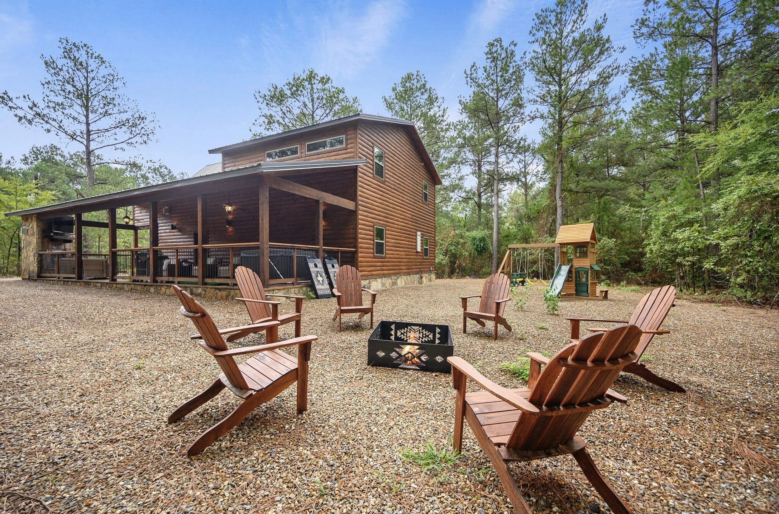 Wooden cabin with a porch in a forested area, surrounded by outdoor furniture and a children's playset with a slide.