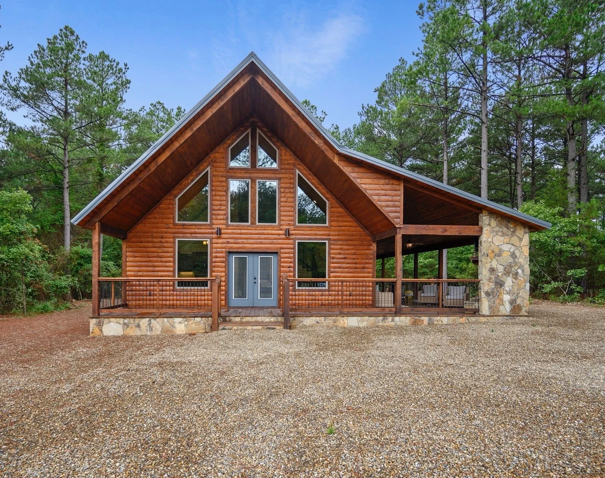 A wooden A-frame house with large triangular windows surrounded by trees, a stone chimney, and a gravel yard.