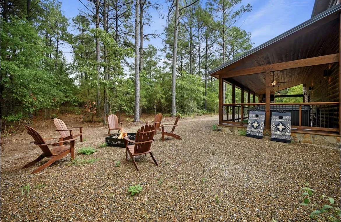 Outdoor patio area with five wooden chairs around a fire pit, surrounded by trees, with a covered deck on the right that has two coolers and a ceiling fan.