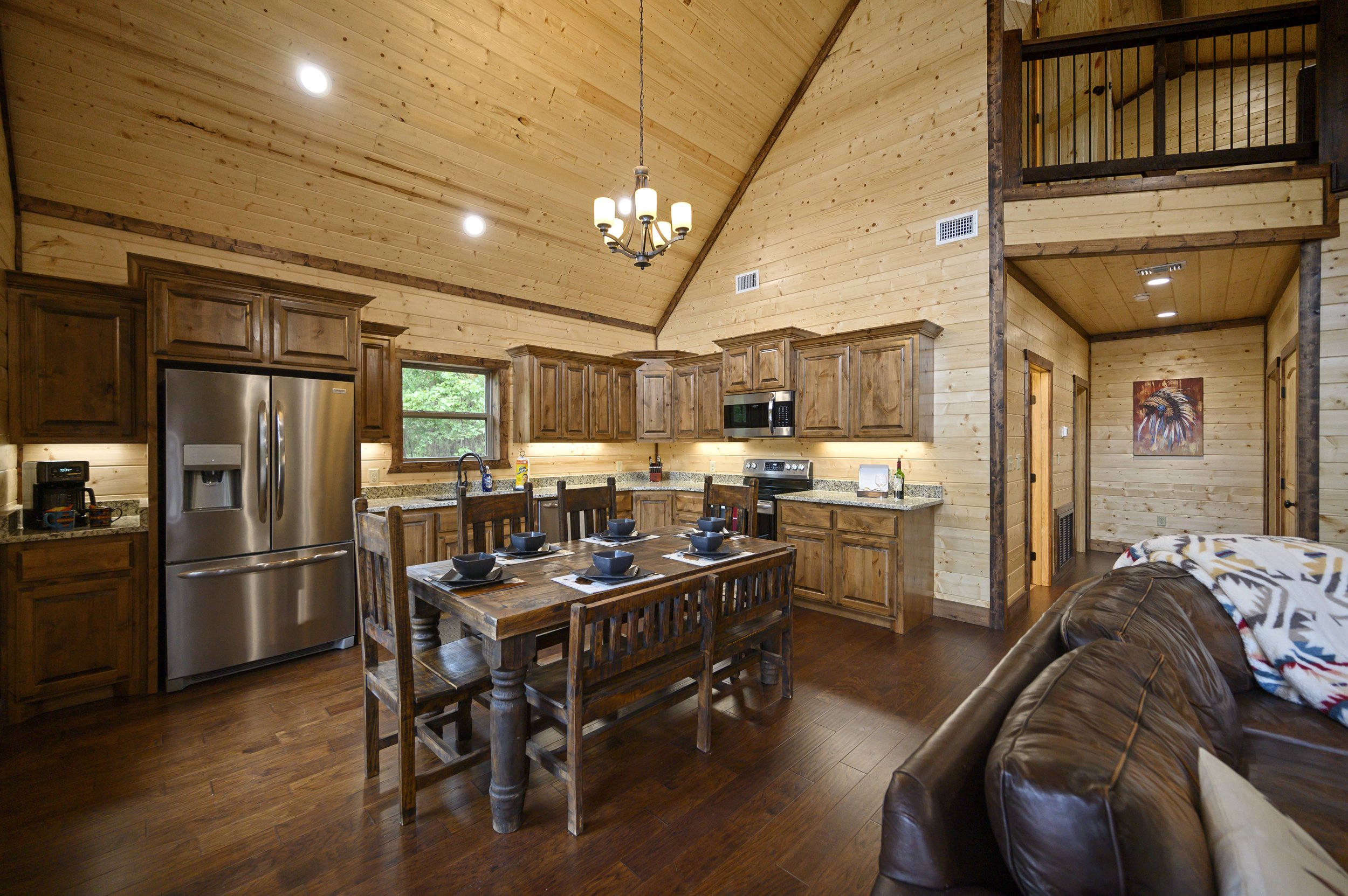 Open-concept kitchen with wooden cabinets, stainless steel refrigerator, granite countertops, dining table with six chairs, and a leather sofa in a cabin-style room with wood-paneled walls and ceiling.