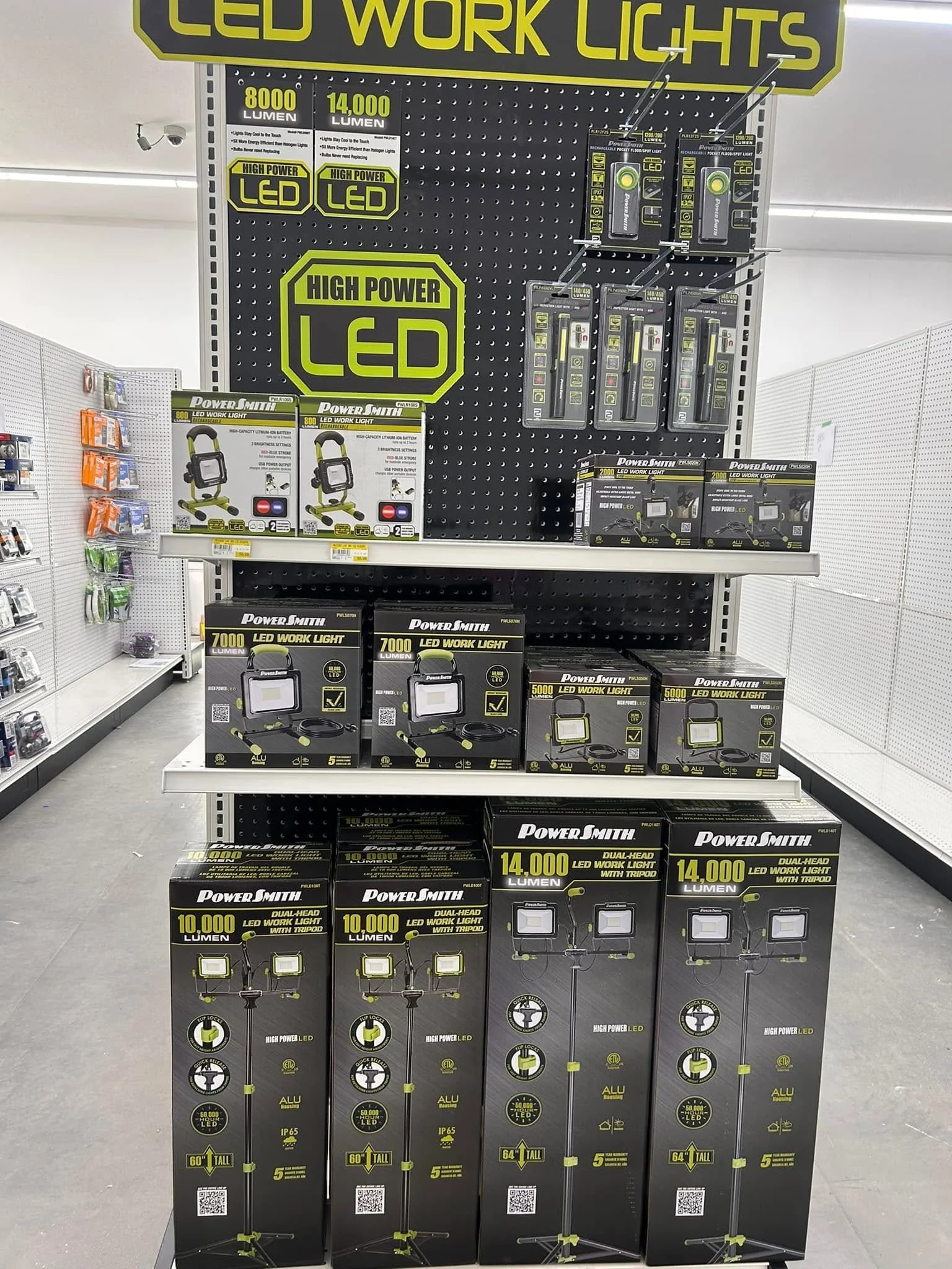 Display of LED work lights and accessories in a retail store. The top sign reads 'LED WORK LIGHTS' in large yellow font. The shelves contain various LED work lights in black and yellow packaging, with specifications such as lumens (ranging from 5,000