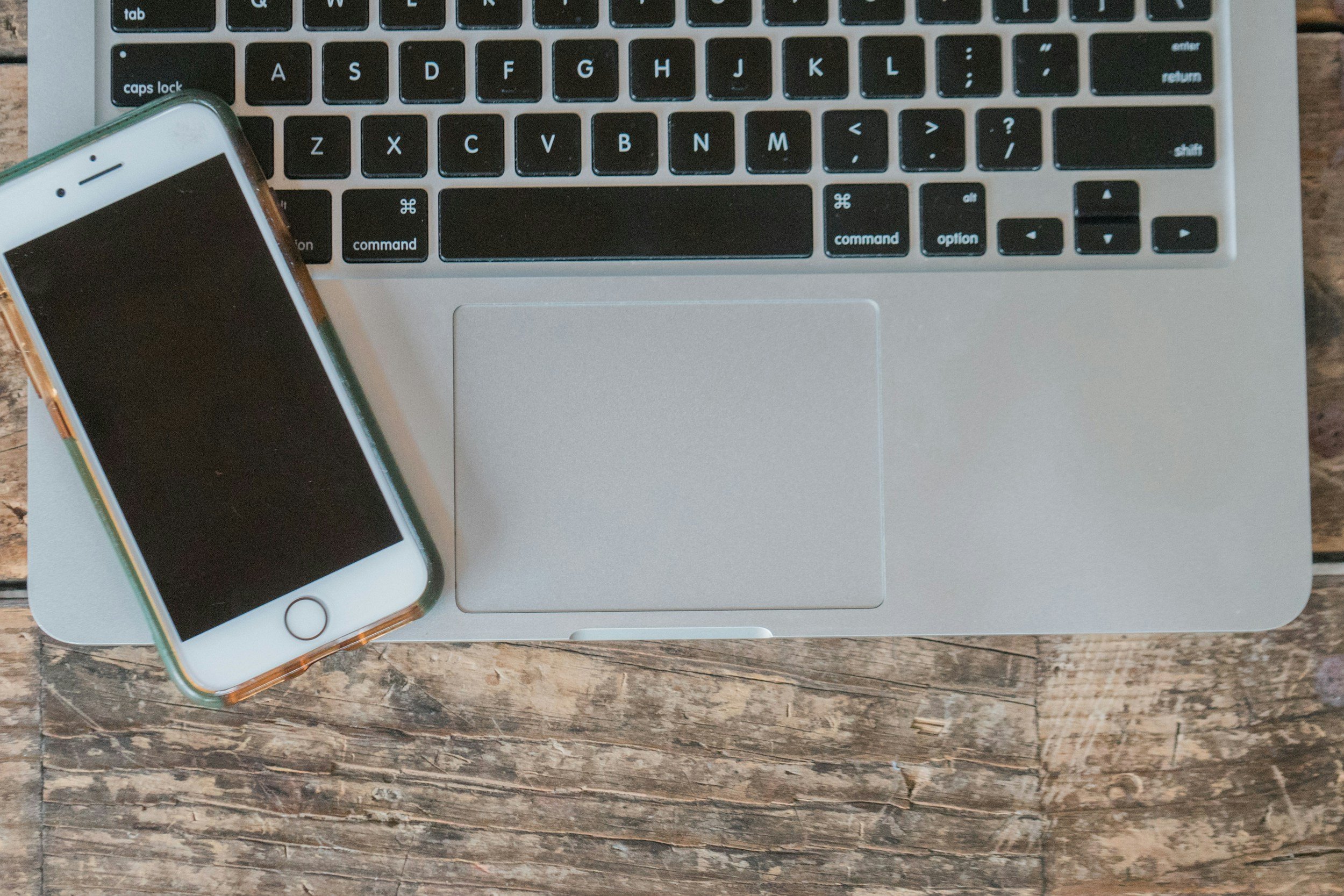 Top-down view of a silver laptop and a white smartphone with a reflective screen on a wooden surface.