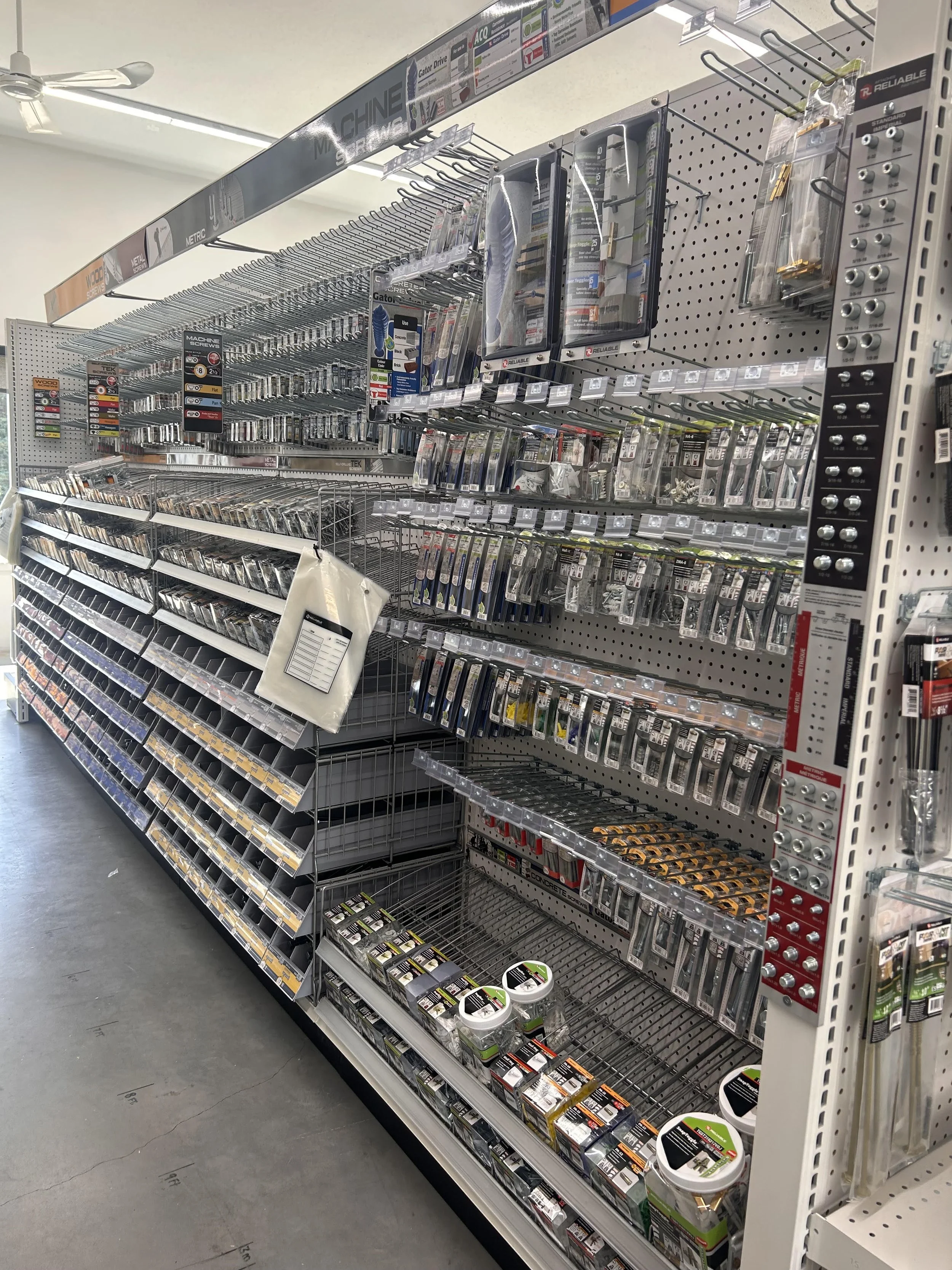 A store aisle featuring a metal rack with screws, hardware, and small tools, with empty shelves and labels for various screw types and sizes.