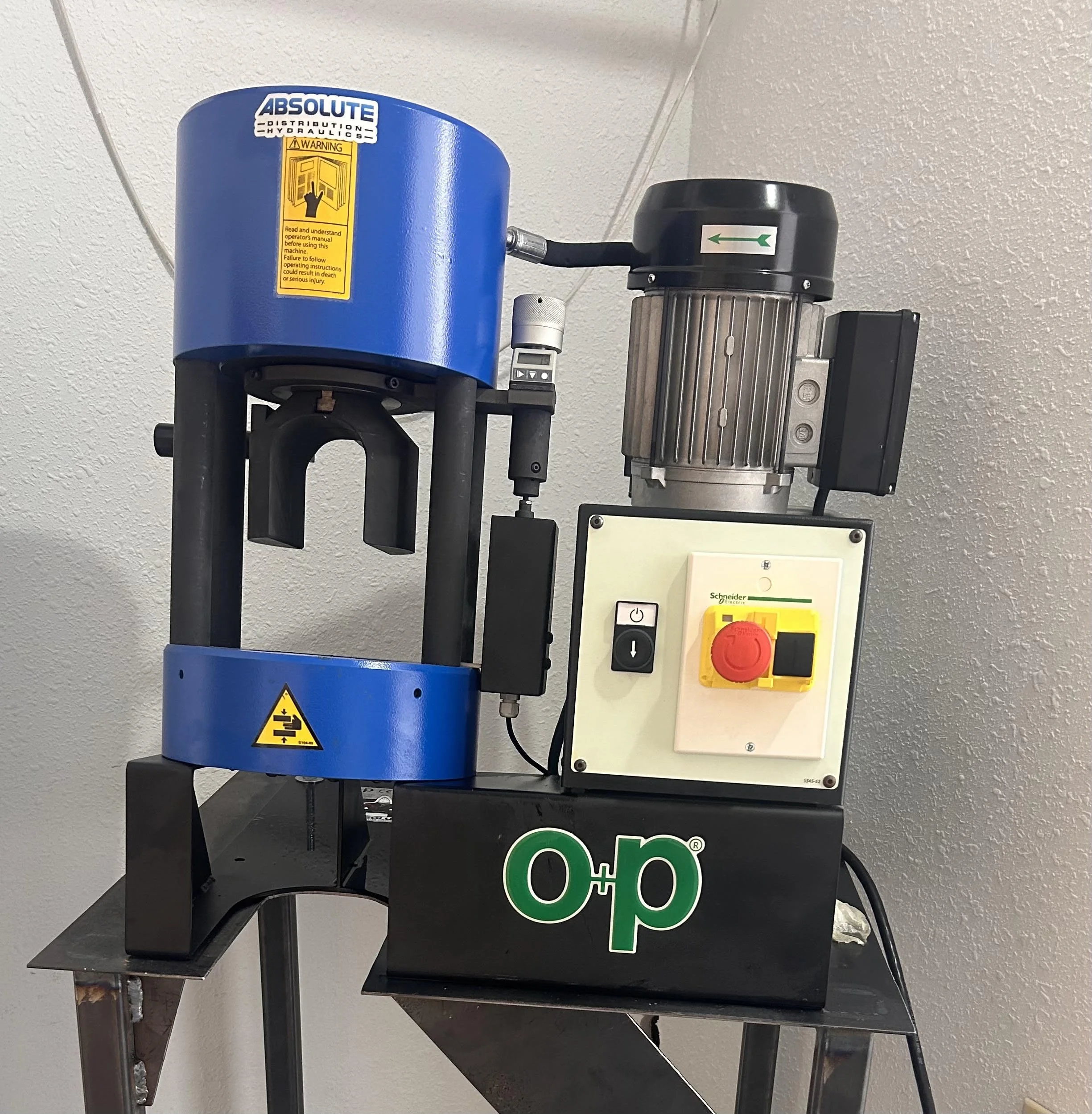 A blue hydraulic machine with a warning label, attached to a control box with buttons and a yellow lock box, in a room with a white textured wall.