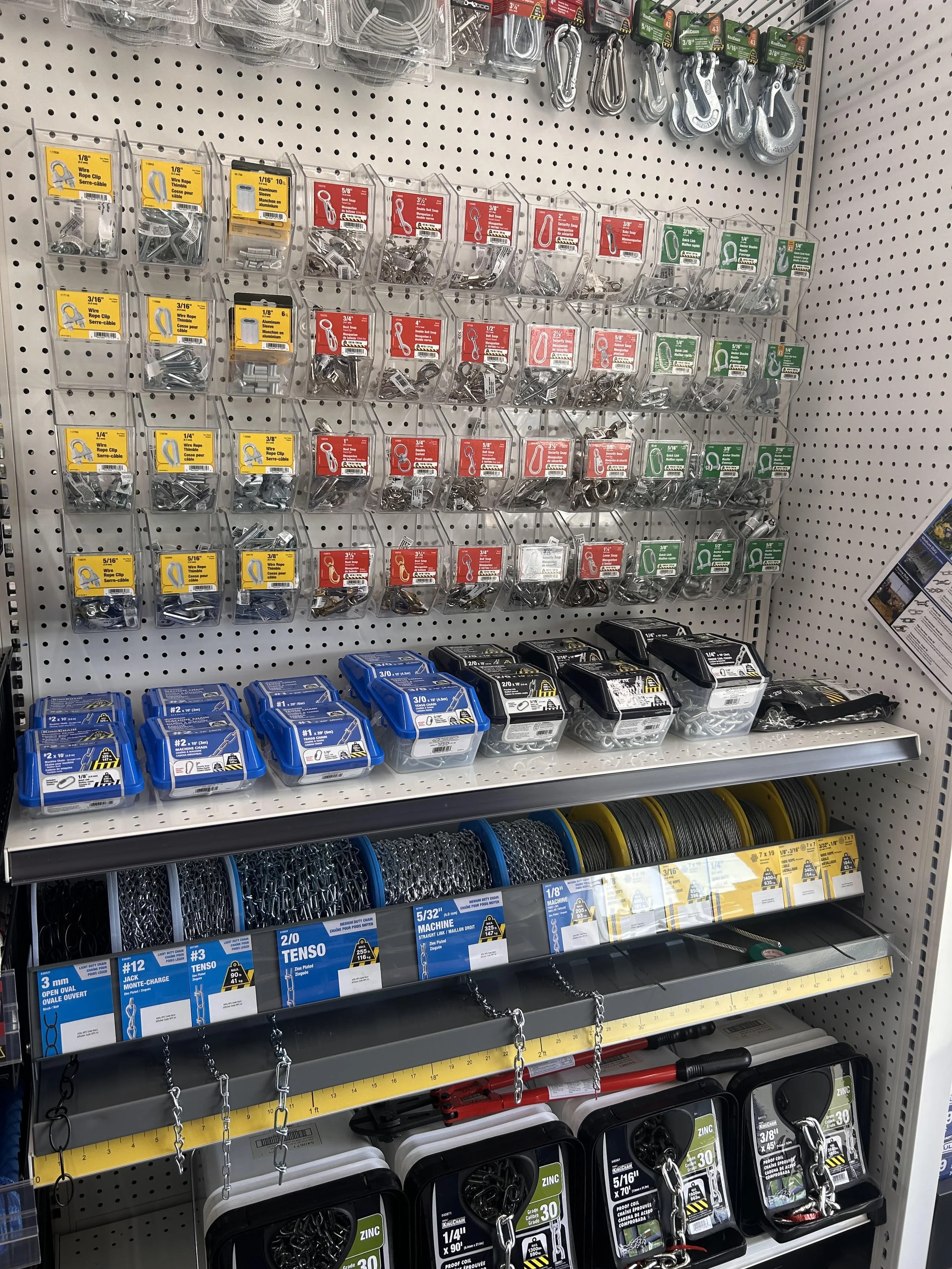 Store shelf displaying various sizes of metal hooks, chains, and fasteners in plastic containers and packaging.