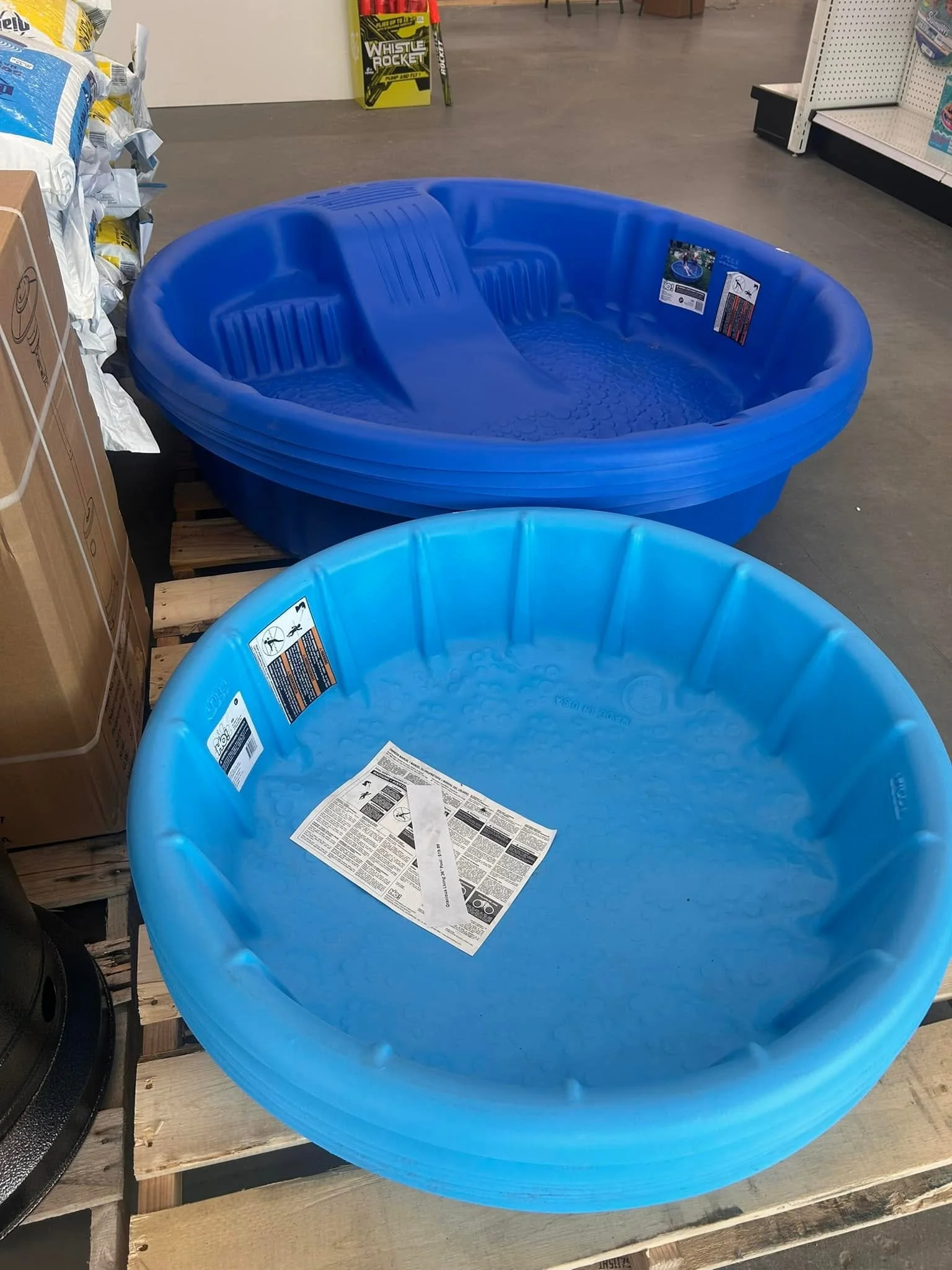 Two blue plastic kiddie pools stacked on a wooden pallet in a store aisle, with instruction labels inside one of the pools.