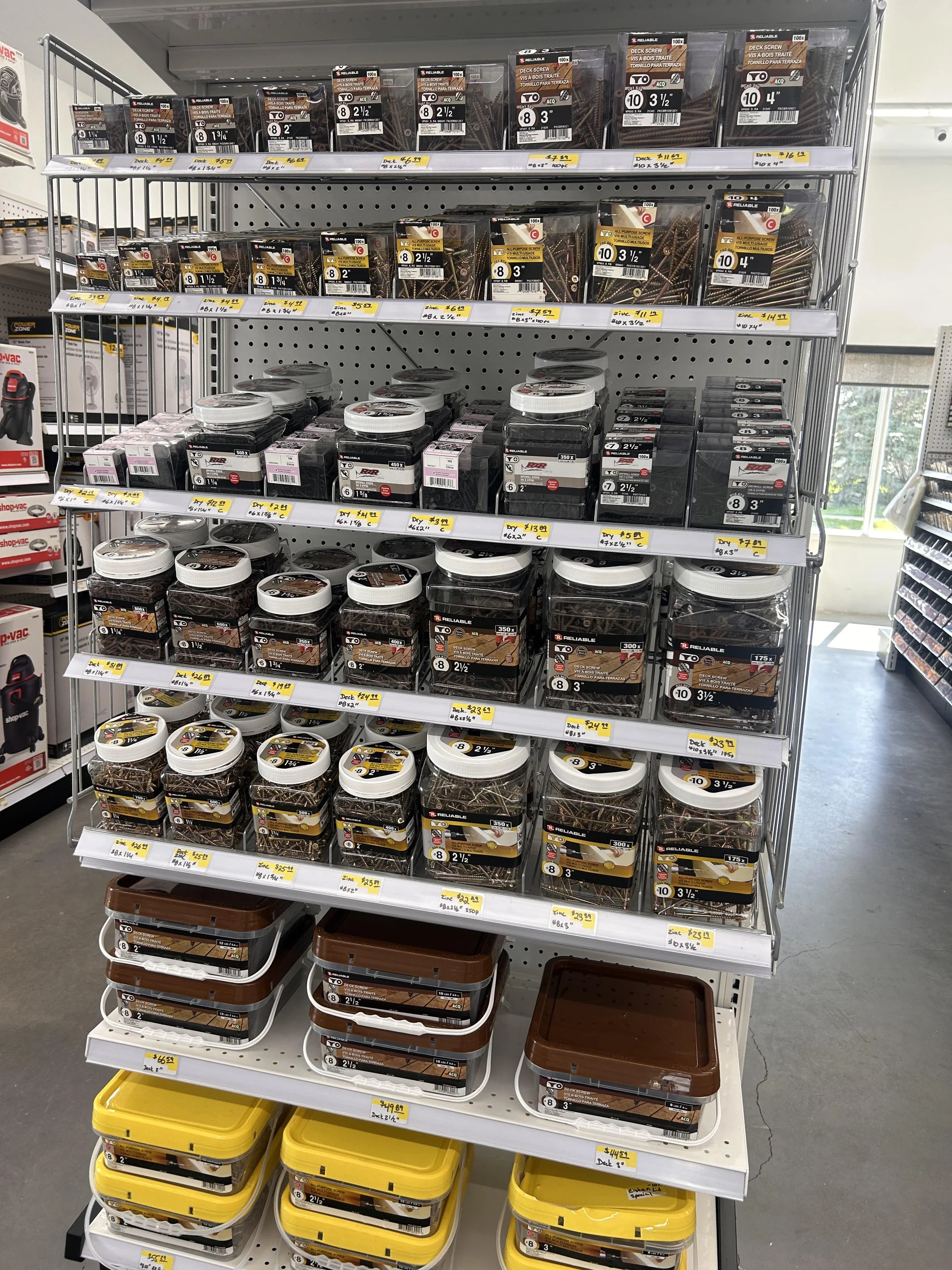 Shelf in hardware store with containers of deck screws arranged by size and color, with price labels.