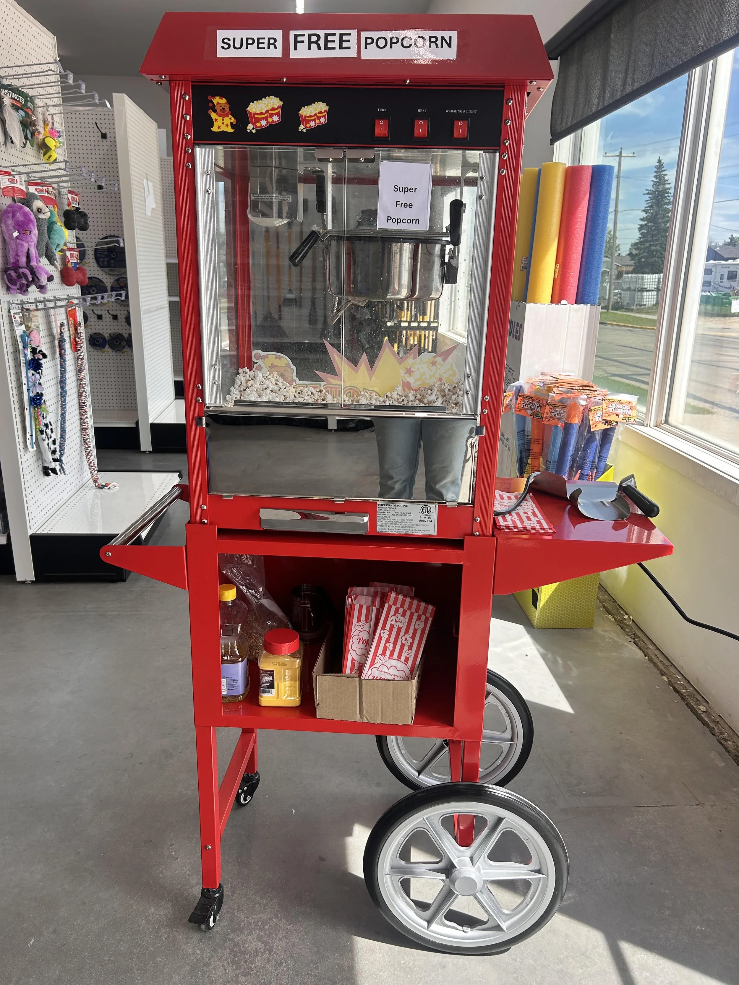 A red popcorn vending machine with a sign that reads "Super Free Popcorn." The machine has a transparent window showing popcorn inside. There are pictures of popcorn and a popcorn character on top of the machine. The machine is located inside a store near a window.
