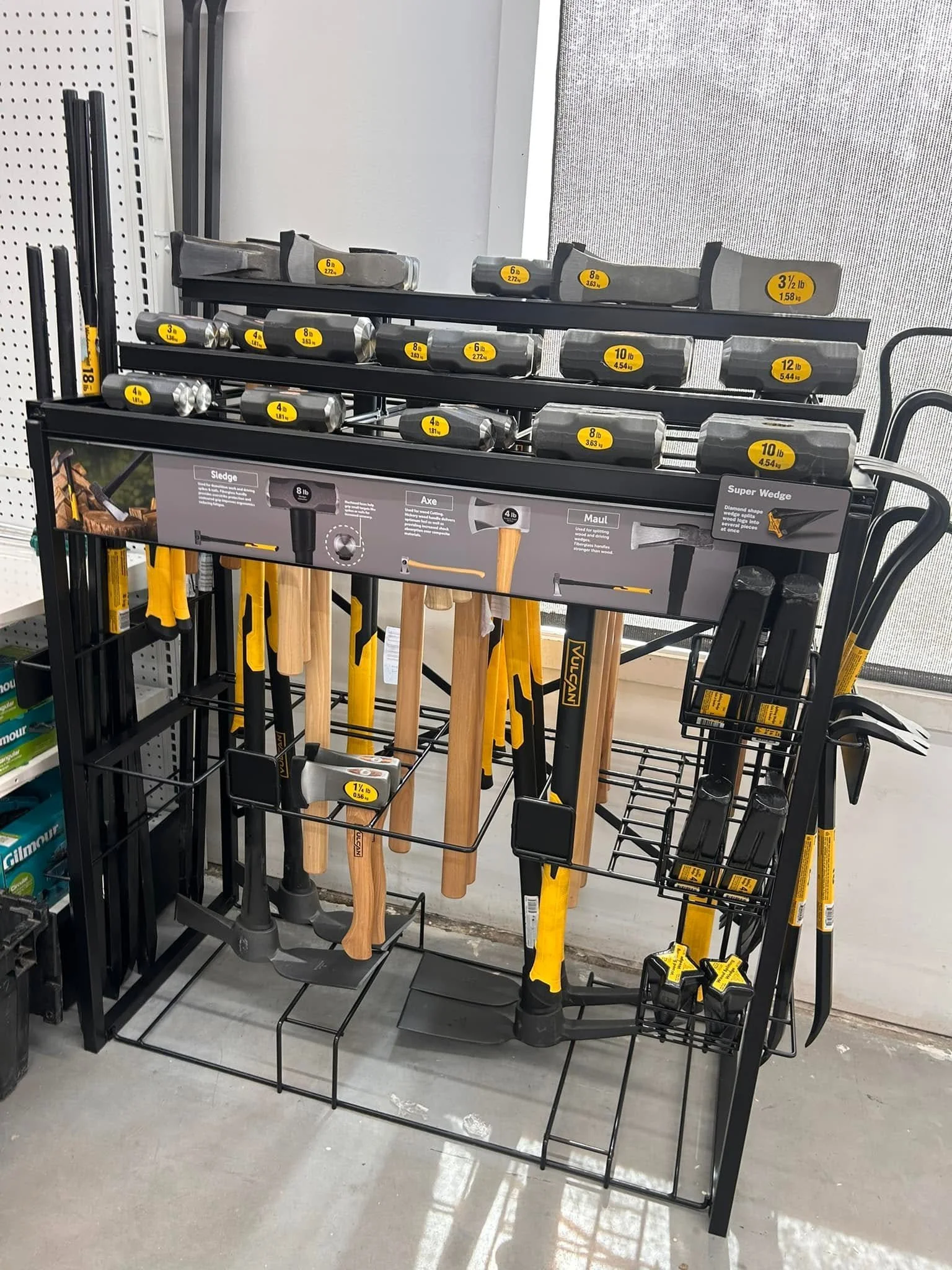 A display of various tools including hammers and axes on a black metal rack inside a store. The tools have yellow and black labels indicating their weights.