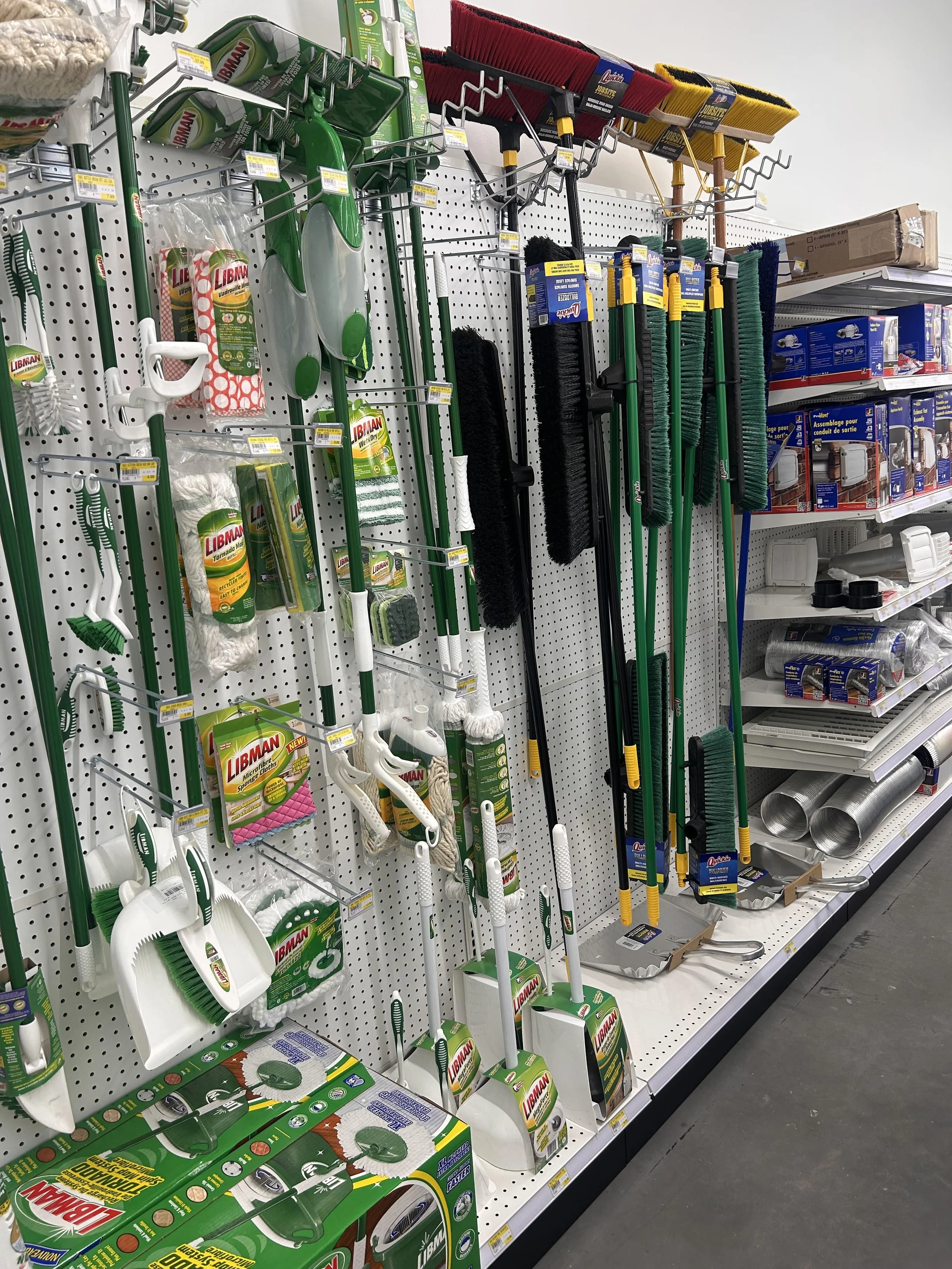 Store shelf with various cleaning tools including brooms, dustpans, and brushes, primarily in green, black, red, and yellow, with some boxes of cleaning supplies nearby.